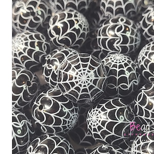 Puede incluir: Un conjunto de cuentas acrílicas negras con un diseño de telaraña blanca. La palabra "ACRYLIC" está escrita verticalmente en letras negras en el lado izquierdo de la imagen. Las cuentas son redondas y brillantes.