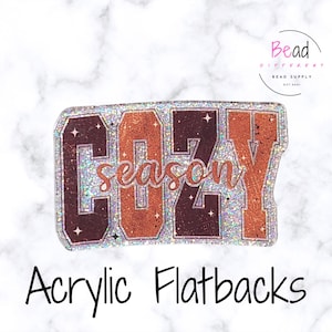 May include: Acrylic flatbacks with the words "COZY season" in brown and orange glitter. The letters are outlined in a silver glitter. The image has a marble background and the words "Bead Different Bead Supply" in the top right corner.