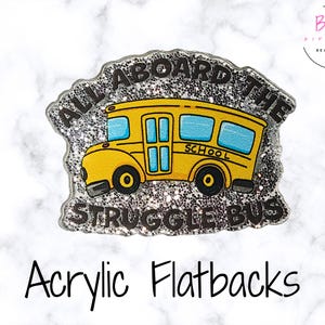 May include: A yellow school bus acrylic flatback with the text "ALL ABOARD THE STRUGGLE BUS". The bus has blue windows and black wheels, set against a glittery silver background. The words "Acrylic Flatbacks" are written below.