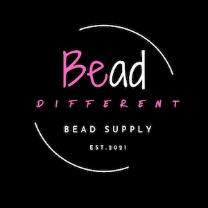 May include: A white circle with a pink handwritten word "Bead" and the text "Bead Different Bead Supply Est. 2021" in black.