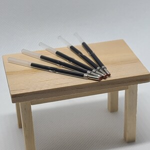 May include: A set of four thin, clear plastic pen ink refills with black ink tubes and metallic silver tips, arranged in a parallel formation on a small, light-colored wooden table with four legs.