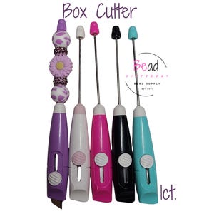 May include: Five box cutters with different colored handles: purple, white, pink, black, and blue. The handles are decorated with beads and a flower. The text "Box Cutter" is at the top of the image. The text "Bead Different Bead Supply" is in a circle at the bottom right of the image.