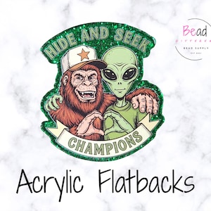 May include: Green glitter acrylic flatback featuring a cartoon Bigfoot and an alien, with the text "HIDE AND SEEK CHAMPIONS." The Bigfoot wears a hat with a star, and the alien wears a green shirt. The words "Acrylic Flatbacks" are at the bottom.