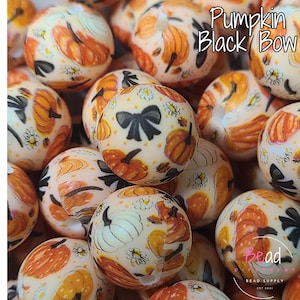 May include: A collection of round beads with a cream background, adorned with orange pumpkins, black bows, and small white flowers. The beads have a glossy finish and are clustered together. The text "Pumpkin Black Bow" is visible.