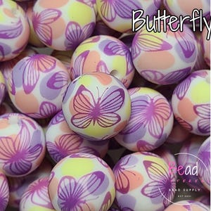 Puede incluir: Primer plano de una colección de cuentas de silicona de colores con un patrón de mariposa. Las cuentas son blancas con acentos rosados, morados y amarillos. Las cuentas están dispuestas en una pila y la palabra "Butterfly" está escrita en letras rosas en la parte superior de la imagen.