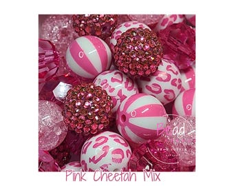 May include: A collection of assorted beads in shades of pink and white. The mix includes round beads with pink cheetah print, pink and white striped beads, translucent faceted pink beads, and round beads covered in small pink rhinestones.