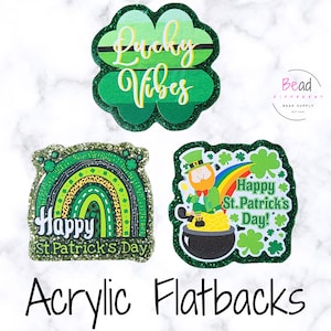 May include: Three glittery acrylic flatbacks with St. Patrick's Day themes. One is a green four-leaf clover with "Lucky Vibes" text. Another features a rainbow and the text "Happy St. Patrick's Day". The third shows a leprechaun and a pot of gold.
