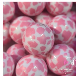 以下が含まれることがあります： 白地にピンクの牛柄模様のアクリルビーズのクローズアップ。ビーズは丸く光沢があり、左側には縦書きで「Acrylic」の文字があります。右下にはBead Supplyのロゴがあります。