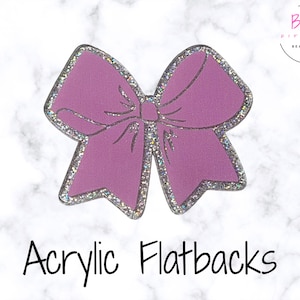May include: A lavender acrylic flatback in the shape of a bow. The bow has a glittery silver outline and a detailed design. The words "Acrylic Flatbacks" are written in black script below the bow.