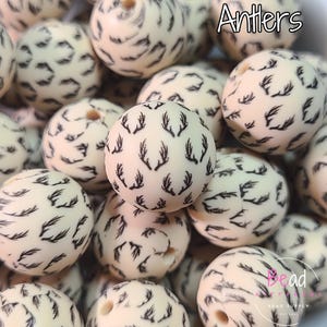 May include: Close-up of ivory-colored beads with black antler patterns. The beads are round and have a smooth texture. The word "Antlers" is written in white text at the top. The beads are for jewelry making and crafts.