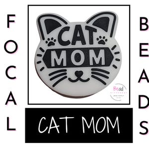 May include: White cat head charm with black outline and the text "CAT MOM" in black lettering. The charm is a focal bead for jewelry making.