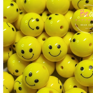 Peut inclure: Gros plan sur un tas de perles acryliques jaunes en forme de smiley. Les perles mesurent environ 2,5 cm de diamètre et ont des yeux et une bouche peints en noir.