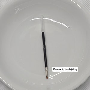 May include: A black ink pen refill with a clear plastic tube at the top and a metal tip at the bottom. A small orange protective cap covers the ballpoint tip. A white label with an arrow points to the tip and reads Remove After Refilling.