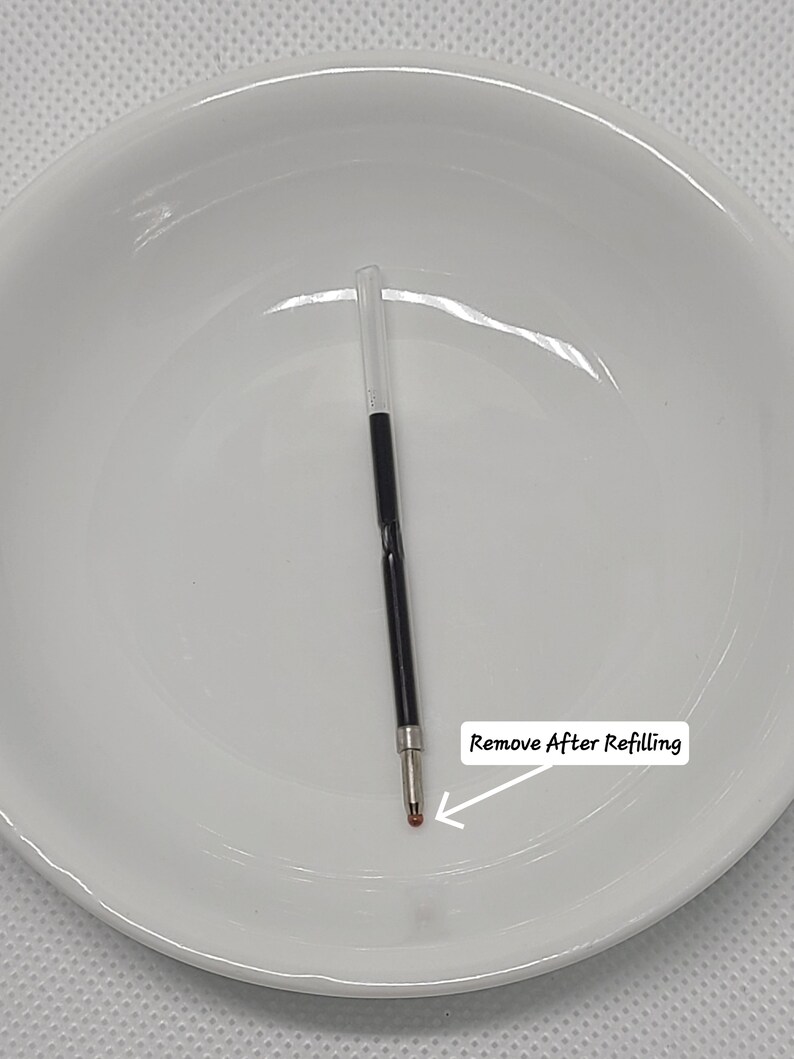 May include: A black ink pen refill with a clear plastic tube at the top and a metal tip at the bottom. A small orange protective cap covers the ballpoint tip. A white label with an arrow points to the tip and reads Remove After Refilling.
