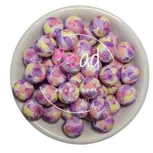 May include: A bowl of colorful silicone beads with a butterfly pattern. The beads are pink, purple, and yellow with white accents. The beads are in a white bowl with the text "Bead Supply Est 2021" on the bottom.