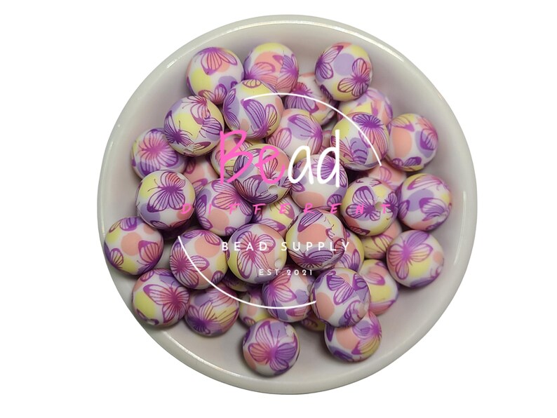 May include: A bowl of colorful silicone beads with a butterfly pattern. The beads are pink, purple, and yellow with white accents. The beads are in a white bowl with the text "Bead Supply Est 2021" on the bottom.