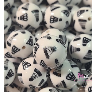May include: White acrylic beads with a glossy finish, each adorned with a black badminton shuttlecock design. The beads are round and have a hole for stringing. The word "Acrylic" is written vertically in black letters on the left.
