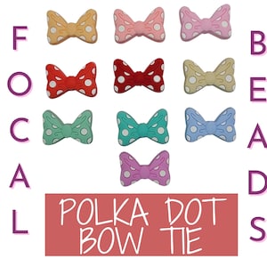 Può includere: Dieci diverse perline di silicone a forma di fiocco con pois in vari colori, tra cui rosa, rosso, giallo, blu e verde. Le perline sono disposte in una griglia con le lettere "FOCAL" a sinistra e "BEADS" a destra. Il testo "POLKA DOT BOW TIE" è visualizzato in un rettangolo rosso in basso.