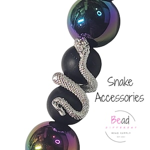 May include: A close-up of a beaded accessory featuring a silver snake wrapped around black and iridescent purple beads. The text "Snake Accessories" is displayed in black, with the Bead Different logo below.
