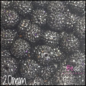 May include: Close-up of a pile of black, sparkly beads. Each bead is approximately 20mm in diameter and covered in small, reflective crystals. The beads have a textured surface and a hole for stringing. The overall effect is a dark, glittering display.