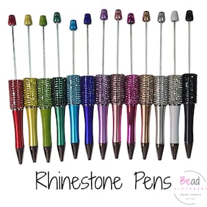 May include: A collection of rhinestone pens in various colors, including red, yellow, green, blue, purple, pink, silver, and black. Each pen features a sparkling rhinestone barrel and a coordinating colored tip. The pens are arranged in a row against a white background.