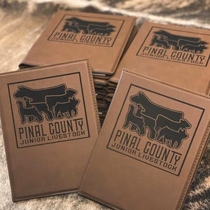 May include: A stack of rectangular brown leatherette folders, each featuring a black laser-engraved logo. The logo depicts silhouettes of a cow, a sheep, and a pig above the text PINAL COUNTY JUNIOR LIVESTOCK, enclosed within a thin black border.