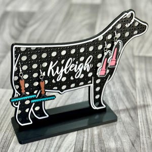 Op de afbeelding: Zwarte koe silhouet oorbellenhouder met witte stippen. De houder heeft de naam "Kyleigh" erop geschreven. De houder heeft drie paar oorbellen die eraan hangen. Een paar is roze hoge hakken, een paar is bruin met een turquoise ontwerp en het andere paar is bruin met een turquoise ontwerp.