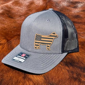May include: A grey trucker hat with a black mesh back and a structured front panel. A tan leather patch on the front displays a cow silhouette filled with an American flag pattern. A black and white sticker on the brim reads Richardson 112.