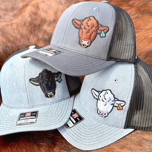 Puede incluir: Tres gorras tipo trucker con parches bordados de cabezas de vaca con anillos nasales de metal. Las gorras tienen paneles frontales grises, parte trasera de malla negra y viseras negras. Incluyen una vaca marrón, una negra y una blanca con etiquetas.