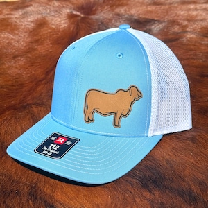 May include: A light blue baseball cap with a white mesh back and a brown leather-like patch featuring a cow silhouette on the front. The brim has a black sticker with the text R 112 The Original MD-LG, featuring a structured crown and eyelets.