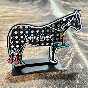 May include: A black and white horse-shaped earring holder with a perforated body and a flat base. The name Kelsey Grace is printed in white script on the side. Two pairs of dangling earrings are attached to the stand, featuring boot and rake designs.