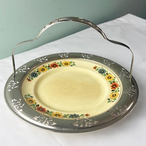 May include: A vintage serving plate with a silver-colored metal handle and rim. The plate features a yellow center with a floral pattern in red, yellow, blue, and orange. The rim has a decorative cut-out design.