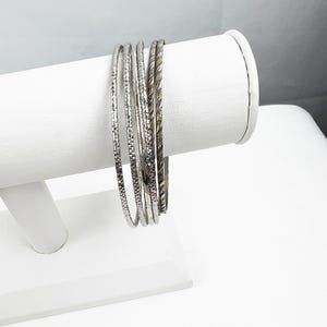 May include: A collection of silver bangles with various textured designs, displayed on a white jewelry stand. The bangles have different patterns, including etched lines and raised textures. The jewelry is presented against a bright, neutral background.