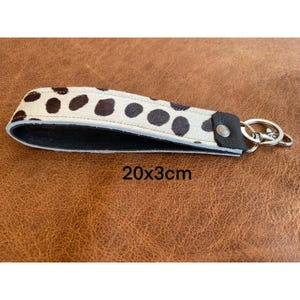 Könnte beinhalten: Ein weißer und schwarzer gepunkteter Leder-Schlüsselanhänger mit einem schwarzen Lederband und einem silbernen Schlüsselring. Der Schlüsselanhänger misst 20 cm x 3 cm.