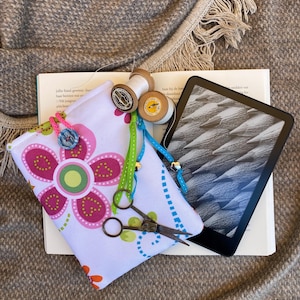 Può includere: Una pochette in tessuto bianco con un motivo a fiore rosa, un nastro blu e verde e un paio di forbici. La pochette è appoggiata su un libro con un e-reader nero sopra il libro. Il libro ha del testo in olandese.
