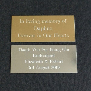 May include: Two rectangular metal plaques. The top plaque is gold-coloured and reads "In loving memory of Daphne Forever in Our Hearts." The bottom plaque is silver and reads "Thank You For Being Our Bridesmaid Elizabeth & Robert 3rd August 2019."