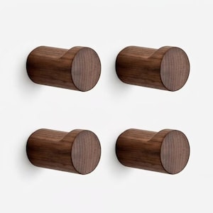 May include: A set of four cylindrical wall hooks made of dark brown wood with a visible grain pattern. Each hook features a flat circular front face and a curved notch on the top side, designed for mounting to a wall surface for hanging items.