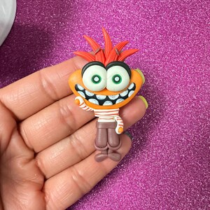 May include: A whimsical orange figurine with large green eyes, a wide grin, and red spiky hair. The figurine wears a striped shirt and brown pants. The figure is a cartoon character.