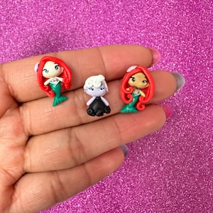 May include: Three small clay figurines of characters from the Little Mermaid. The figurines are red, purple, and green. The figurines are on a pink sparkly background.
