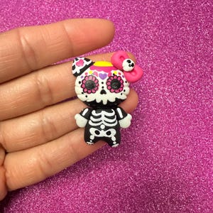 May include: A Day of the Dead themed figurine in the shape of a bear. The bear is white with a black skeleton body and a skull face with pink flower accents. It has a pink bow and is set against a sparkly pink background.