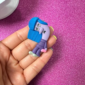 May include: A handmade polymer clay figurine of a cartoon character with a blue fringe hairstyle, large eyes, and a purple body. The character wears a purple outfit and holds a pink object. The figurine is held in a hand against a sparkly pink background.