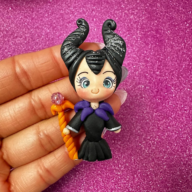 May include: A handmade figurine of a character with black horns, a black dress, and a purple collar. The figurine holds an orange staff with a pink crystal. The character has large blue eyes against a pink background.