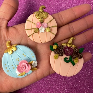 May include: Three decorative pumpkin ornaments with different designs. One is light blue with gold accents and a pink rose, another is pink with gold accents and pink and white flowers, and the last is pink with gold accents and brown and burgundy flowers.