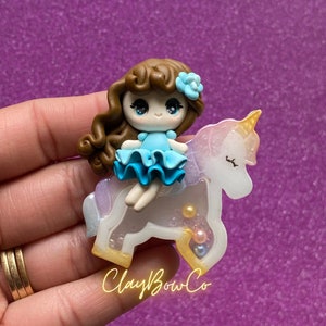May include: A brown-haired girl doll wearing a blue dress sits on a white unicorn figurine with iridescent glitter accents. The unicorn has a gold horn and closed eyes. The figurine has a clear resin base with iridescent glitter and small pearl-like beads.