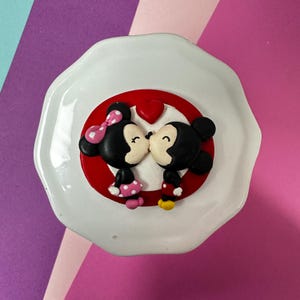 May include: A white, irregularly shaped dish featuring a red circle with Mickey and Minnie Mouse kissing, and a red heart above them. Minnie wears a pink bow and polka-dot skirt. Mickey wears red shorts. The background is pink and purple.