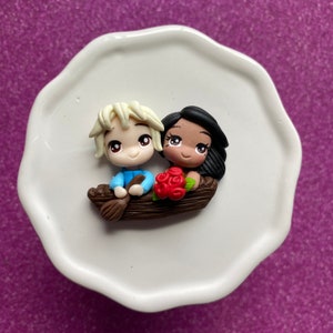 May include: A handcrafted polymer clay figurine depicts a couple in a boat, with the man in a blue shirt and the woman with black hair holding red roses. The boat is brown, and the figurine sits on a white, scalloped plate.