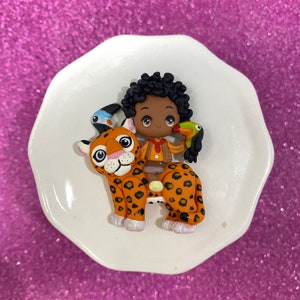 May include: A brown and black jaguar figurine with a brown-skinned person figurine riding on its back. The person figurine is wearing a brown shirt and a red bandana. A blue and green toucan figurine is perched on the person's shoulder.