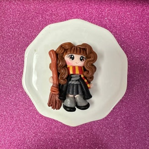 May include: A brown-haired girl doll with a black dress and a red and yellow scarf holds a brown broom. The doll is on a white plate.