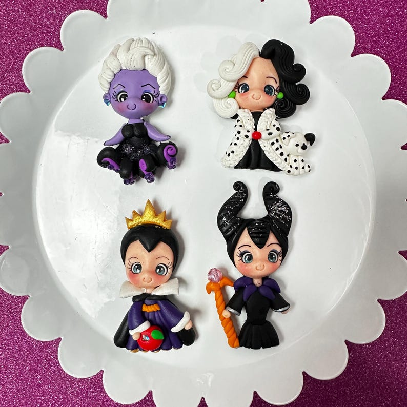 May include: Four miniature figurines of Disney villains, including Ursula, Cruella de Vil, the Evil Queen, and Maleficent, are arranged on a white, scalloped plate. Each figure is brightly coloured and detailed, showcasing their iconic looks. The plate sits on a pink glitter surface.