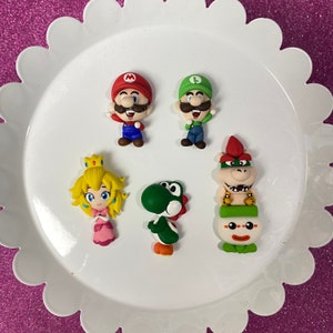 May include: A collection of colorful, miniature figurines of characters from the Super Mario video game series. The figurines include Mario, Luigi, Princess Peach, Yoshi, and Bowser Jr. They are arranged on a white, scalloped-edge plate.